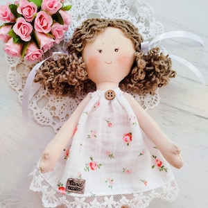 May include: A handmade doll with brown curly hair, wearing a white dress with pink floral print and a lace trim. The doll is holding a bouquet of pink roses and is sitting on a white crocheted doily. The dress has a small brown tag that says "Hand Made".