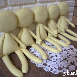 May include: Five unpainted fabric dolls, all the same size and shape, are arranged in a row on a white lace doily. The dolls are made of a soft, light yellow fabric and have no facial features.