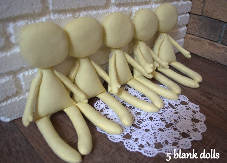 May include: Five unpainted fabric dolls, all the same size and shape, are arranged in a row on a white lace doily. The dolls are made of a soft, light yellow fabric and have no facial features.