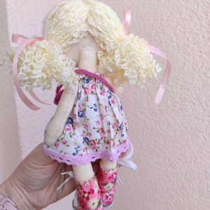 May include: A handmade doll with blonde yarn hair styled in pigtails, adorned with pink ribbons. The doll wears a floral print dress with a pink lace trim and matching pink floral shoes. The doll is held in a person's hand.