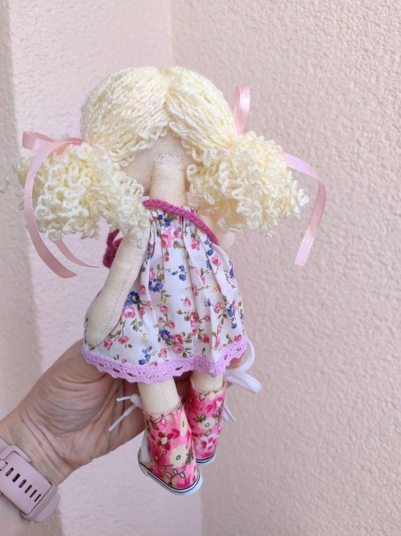 May include: A handmade doll with blonde yarn hair styled in pigtails, adorned with pink ribbons. The doll wears a floral print dress with a pink lace trim and matching pink floral shoes. The doll is held in a person's hand.