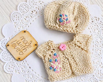 May include: A cream-colored knitted baby set, including a beret and a cardigan, adorned with pink and blue floral embroidery. The cardigan has a pink button closure. A wooden tag with the words "made with love" is also present.