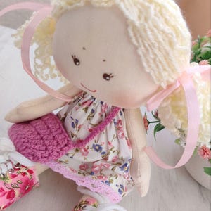 May include: A handmade doll with blonde yarn hair tied with pink ribbons. The doll wears a floral print dress with a pink crocheted purse. The doll's shoes are also decorated with a floral pattern.