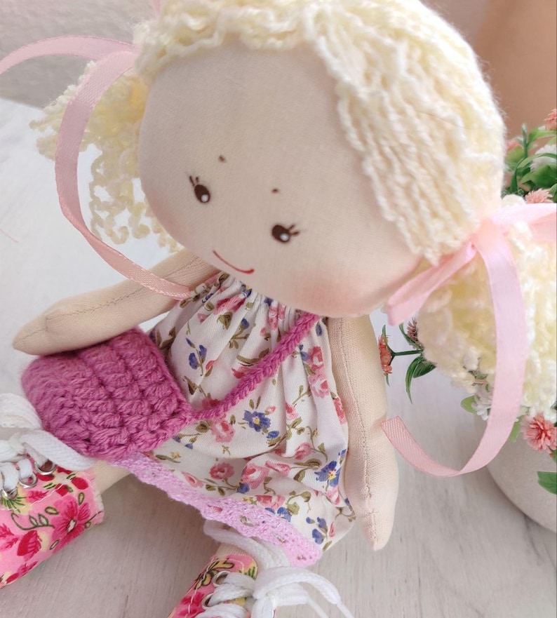May include: A handmade doll with blonde yarn hair tied with pink ribbons. The doll wears a floral print dress with a pink crocheted purse. The doll's shoes are also decorated with a floral pattern.
