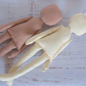 May include: Two unstuffed fabric dolls, one beige and one cream, lying on a white wooden surface. The cream doll has a white lace headband with pink and green flowers.