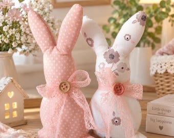 May include: Two stuffed fabric rabbit figures. One is pink with white polka dots, the other is white with a brown floral pattern. Each rabbit features a pink lace ribbon tied around its neck with a small wooden button at the center.
