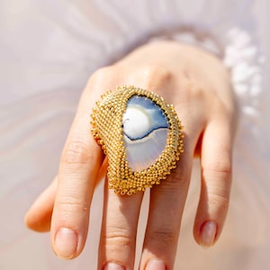 Könnte beinhalten: Ein auffälliger Ring mit einem großen, halbkreisförmigen, polierten weiß-blauen Stein-Cabochon. Der Stein ist von einem dichten, strukturierten Rand aus winzigen, metallisch goldenen Perlen umgeben, die einen schimmernden Rahmen bilden.