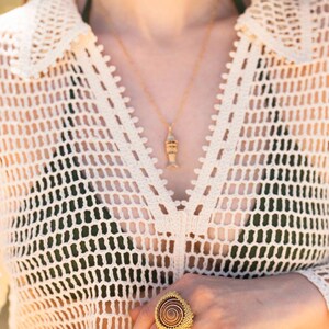 Puede incluir: Un collar dorado con un peque&ntilde;o colgante en forma de pez y un anillo grande con un dise&ntilde;o de espiral marr&oacute;n rodeado de intrincadas cuentas doradas, usado con una blusa de crochet color crema sobre una prenda de color verde oscuro.
