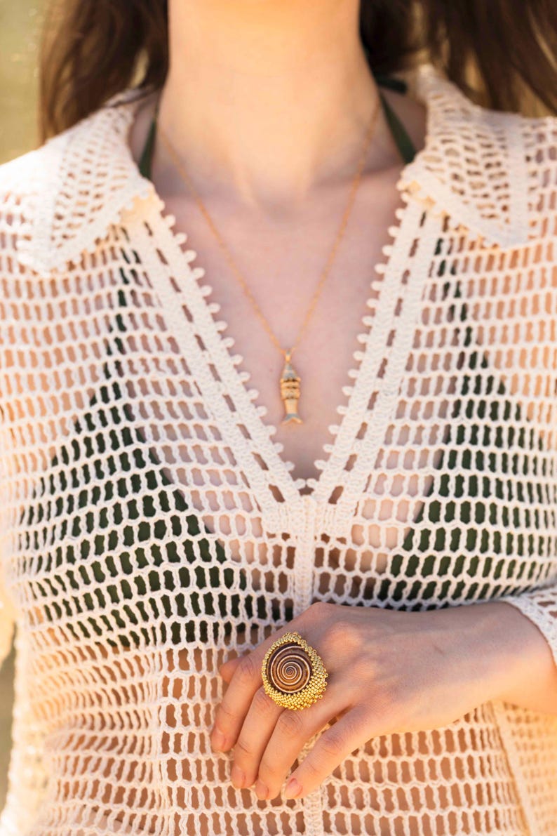 Puede incluir: Un collar dorado con un peque&ntilde;o colgante en forma de pez y un anillo grande con un dise&ntilde;o de espiral marr&oacute;n rodeado de intrincadas cuentas doradas, usado con una blusa de crochet color crema sobre una prenda de color verde oscuro.