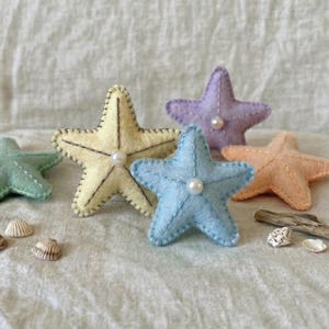 May include: A collection of six felt starfish in various pastel colors: green, yellow, blue, purple, and peach. Each starfish is embellished with a pearl in the center and stitched edges. Seashells and driftwood are scattered around the starfish.