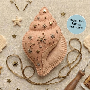May include: A light pink felt seashell with embroidered details and star embellishments. The image includes a digital pattern label, gold stars, and crafting tools. The seashell is surrounded by other felt sea-themed shapes.