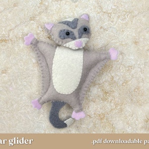Puede incluir: Un juguete de peluche de azúcar planeador de fieltro con pelaje gris, vientre blanco y orejas y patas rosas. El juguete está acostado sobre una superficie texturizada marrón claro. "sugar glider" y ".pdf downloadable pattern" están escritos en las esquinas inferiores de la imagen.