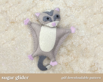 May include: A felt sugar glider plush toy with gray fur, a white belly, and pink ears and paws. The toy is lying on a light brown textured surface. "sugar glider" and ".pdf downloadable pattern" are written in the bottom corners of the image.