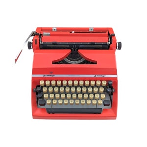 May include: A bright red Scheidegger International typewriter. The typewriter has a standard QWERTY keyboard with off-white keys. The text "Scheidegger International" is printed on the front. The typewriter is a vintage office supply.