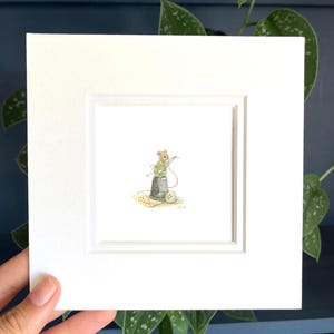 Puede incluir: Una impresión en acuarela enmarcada que presenta una ilustración caprichosa de un ratón cosiendo con un dedal y una aguja. La obra de arte incluye un carrete de hilo, un botón y un trozo de tela. La impresión está enmarcada en blanco y sostenida por una mano.
