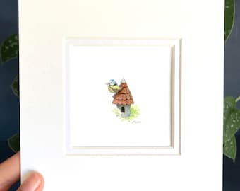 May include: A watercolor painting of a bluebird perched on a small birdhouse with a brown roof. The birdhouse is surrounded by green foliage.
