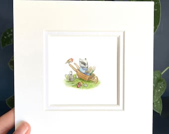 May include: A watercolor painting of a badger sitting in a wheelbarrow reading a book. A robin perches on the wheelbarrow handle. The scene is set in a garden with green grass and red berries.