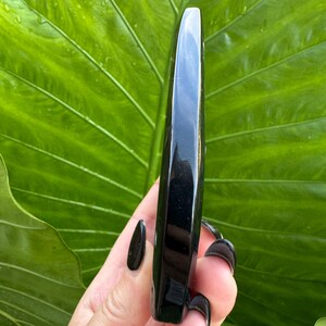 May include: A polished, black obsidian wand. The wand is held against a backdrop of a large, green leaf. The wand is smooth and rectangular with a rounded tip. The obsidian has a glossy, reflective surface.
