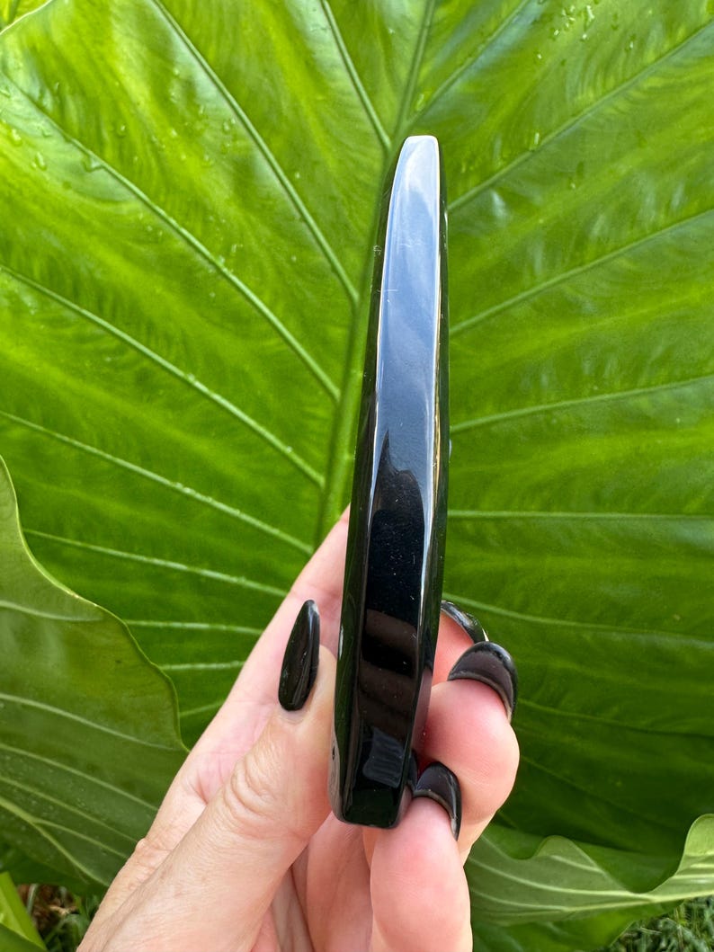 May include: A polished, black obsidian wand. The wand is held against a backdrop of a large, green leaf. The wand is smooth and rectangular with a rounded tip. The obsidian has a glossy, reflective surface.