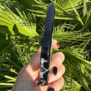 May include: A polished, black and white agate wand. The wand is rectangular with rounded edges and a smooth, glossy finish. The stone has a marbled pattern of black and white. The background is a lush green foliage.