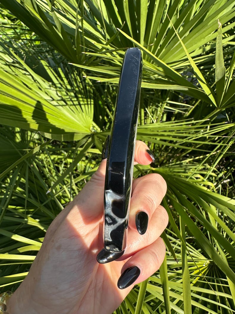 May include: A polished, black and white agate wand. The wand is rectangular with rounded edges and a smooth, glossy finish. The stone has a marbled pattern of black and white. The background is a lush green foliage.