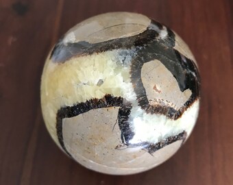 May include: A polished sphere of septarian stone, a type of sedimentary rock with a unique pattern of brown, black, and cream colored bands. The sphere is smooth and has a glossy finish.