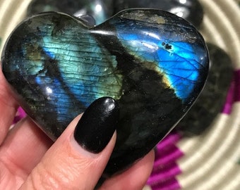 May include: Three heart-shaped labradorite crystals, one of which is being held in a hand. The crystals are a dark blue-green color with iridescent flashes of color.