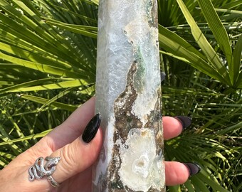 May include: A tall, polished crystal obelisk with a pointed top. The crystal features a mix of white, brown, and green hues, with visible patterns and inclusions. The obelisk is held against a backdrop of green foliage.