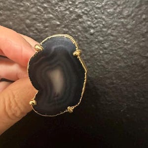 May include: A ring featuring a large, irregular, dark grey and black agate slice with natural concentric banding. The stone is set in a gold-toned metal prong setting that follows the organic, uneven shape of the agate slice, highlighting its unique texture.