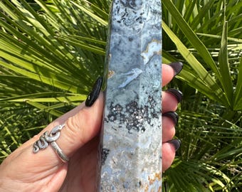 May include: A tall, rectangular crystal obelisk with a pointed top. The crystal features a mix of blue, white, gray, and brown colors, with a marbled appearance. The obelisk is held against a backdrop of green foliage.