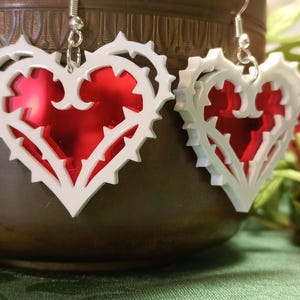 May include: A pair of white heart-shaped earrings with a red heart design inside. The earrings have a delicate, intricate design with a thorny vine pattern.
