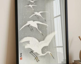 Op de afbeelding: Een ingelijste kunstdruk met een groep witte kraanvogels die vliegen tegen een grijze achtergrond met witte spikkels. Het kunstwerk bevat een rood zegel in de linkerbenedenhoek en is geplaatst in een dunne, zwarte, rechthoekige lijst.