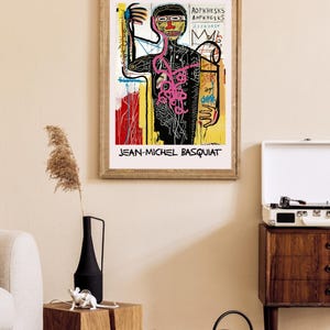 Puede incluir: Impresi&oacute;n enmarcada de una obra de arte de Jean-Michel Basquiat. La obra representa una figura estilizada con l&iacute;neas audaces y colores vibrantes, con el nombre del artista en la parte inferior. La impresi&oacute;n est&aacute; en un marco de madera.