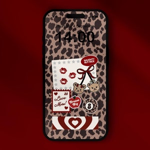 May include: A smartphone with a leopard print case and a red and white heart design on the screen. The screen also features several stickers, including a red heart with the text "Dream Big", a red apple with the text "Love More", and a red ribbon with the text "Naughty But Nice". The time on the screen is 14:00.