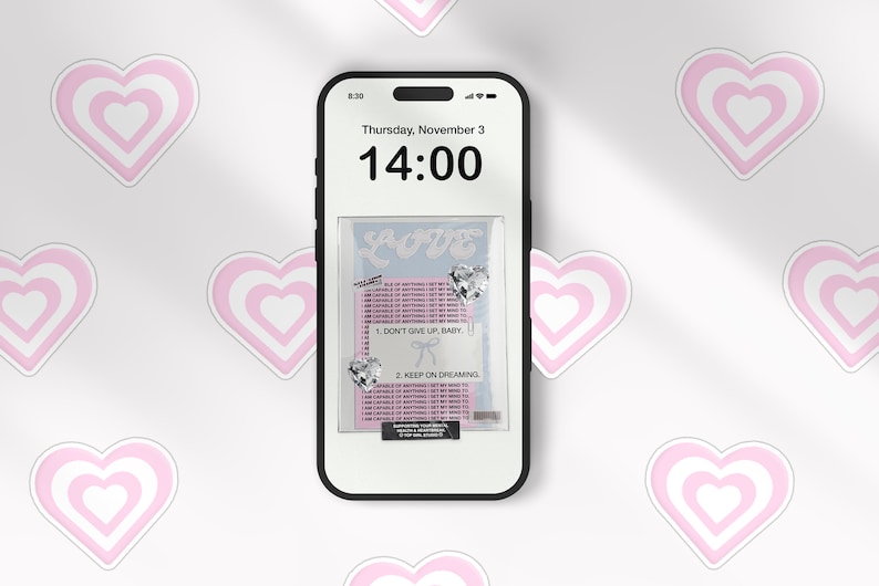 May include: A white smartphone with a black screen displaying the time 14:00 and the date Thursday, November 3. The screen also shows a pink and white heart design with the word "LOVE" and the text "DON'T GIVE UP, BABY" and "KEEP ON DREAMING".