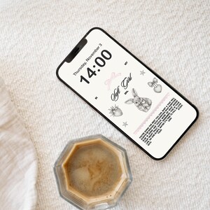May include: A smartphone displaying a digital clock with the time 14:00 and the date Thursday, November 3. The screen also features a pink ribbon, a strawberry, a star, a bunny, and a heart with a bow. The phone is lying on a white textured surface next to a glass cup of coffee.