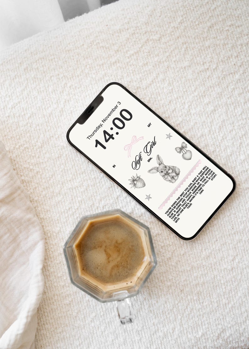 May include: A smartphone displaying a digital clock with the time 14:00 and the date Thursday, November 3. The screen also features a pink ribbon, a strawberry, a star, a bunny, and a heart with a bow. The phone is lying on a white textured surface next to a glass cup of coffee.