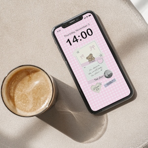 Puede incluir: Un smartphone con un fondo de pantalla de cuadros rosas con un gráfico de osito de peluche y el texto "You deserve the world, baby. Go claim it!". Una taza de café está al lado del teléfono sobre una superficie de tono neutro.