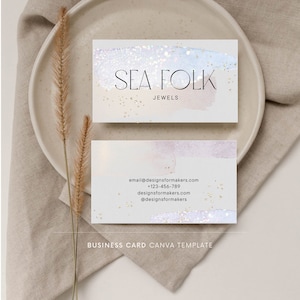Op de afbeelding: Een visitekaartje sjabloon voor een juweliersbedrijf genaamd "Sea Folk Jewels". De kaart heeft een aquarel achtergrond in lichtblauw en roze met gouden glitter accenten. De naam van het bedrijf en de website zijn op de kaart gedrukt.