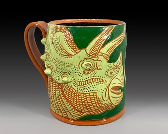 May include: A ceramic mug with a green background and a detailed illustration of a yellow and orange triceratops.