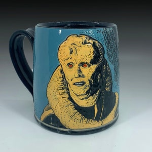 Puede incluir: Una taza de cerámica con asa negra y fondo azul. La taza presenta una ilustración pintada a mano de un personaje con ojos naranjas y un cuello con estampado amarillo y negro.