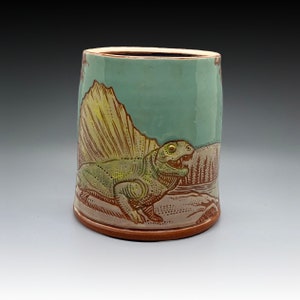 Può includere: Tazza in ceramica con un'illustrazione dettagliata di un dinosauro Dimetrodon. La tazza presenta uno sfondo azzurro con un disegno di dinosauro marrone e verde, e un bordo e una base marroni. Il dinosauro è ambientato in un paesaggio.