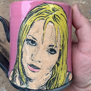 Puede incluir: Una taza de cerámica rosa con asa negra presenta una ilustración pintada a mano de una mujer con cabello rubio. El rostro de la mujer está detallado con contornos negros y puntillismo.