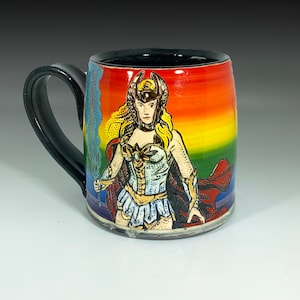 Op de afbeelding: Een keramische mok met een zwart handvat en rand, met een kleurrijk regenboogverloopontwerp. De mok toont een gedetailleerde illustratie van een vrouwelijke krijger in gouden en witte bepantsering, met een rode cape. De mok is een uniek kunstwerk.