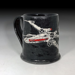 Könnte beinhalten: Schwarze Keramik-Tasse mit Henkel, mit einer weißen und roten Illustration eines Raumjägers. Die Tasse hat eine strukturierte Oberfläche und einen breiten Boden. Der Griff ist schwarz und gebogen.