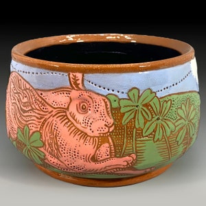May include: A ceramic planter with a hand-painted design of a pink rabbit and green shamrocks. The planter has a brown rim and a light blue background.