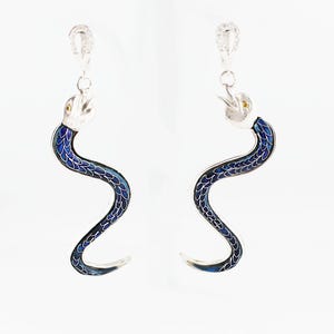 May include: A pair of silver-toned drop earrings featuring a snake design. Each snake has a metallic silver head with small yellow eyes and a long, curved body filled with a vibrant blue enamel scale pattern, suspended from a silver-toned clasp.
