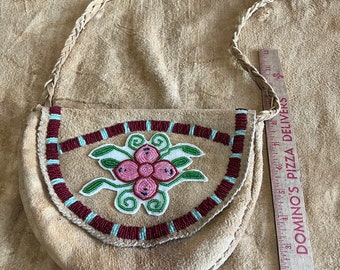 May include: A small, tan leather pouch with a beaded floral design. The pouch has a braided leather strap and is decorated with red, white, and blue beads.