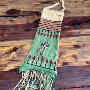 May include: A green and tan leather bag with fringe, painted with geometric designs and red accents. The bag has a strap and a beaded circle with bells attached.