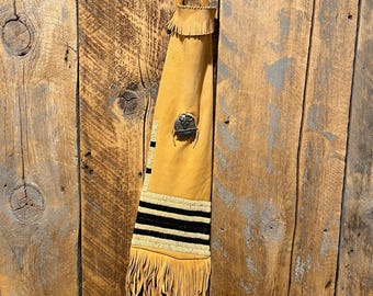 May include: A tan leather bag with black and white trim and fringe. The bag has a decorative metal piece on the front.